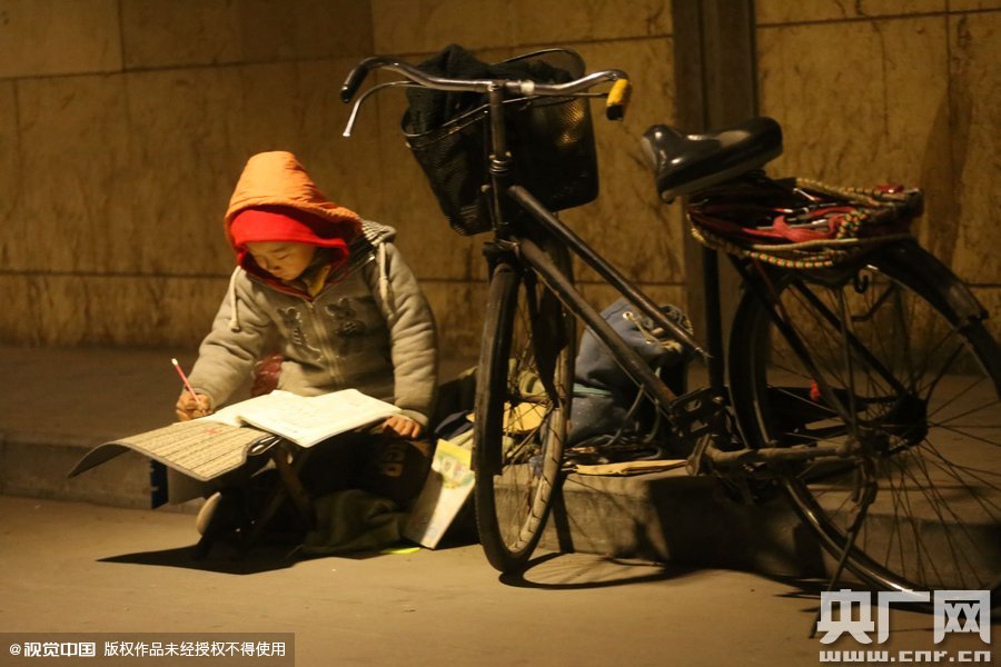 鄭州中原路京廣鐵路大橋下涵洞的馬路牙子邊，一位戴著紅色辣椒帽的小男孩趴在一個(gè)小馬扎上，上面鋪著一張車墊，在微弱的燈光下寫作業(yè)